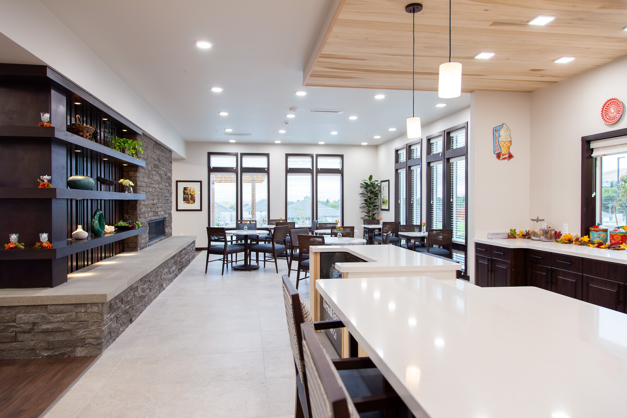 Spacious modern kitchen and dining area with large windows, white countertops, wooden cabinets, pendant lights, and decorative shelves. The room has a bright, open feel with plants and decor accentuating the space.