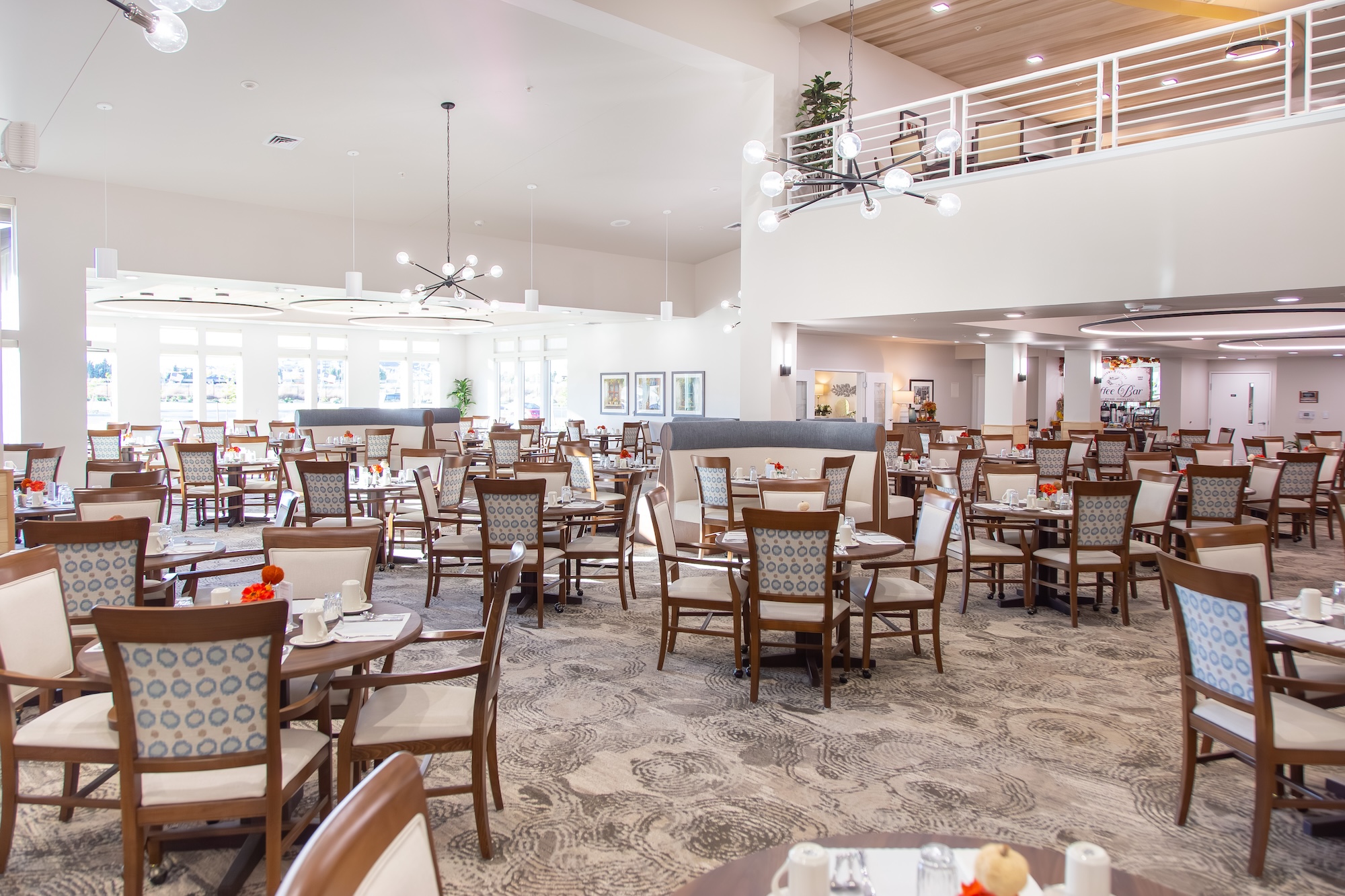 A spacious, well-lit dining area features numerous wooden tables and chairs arranged neatly. Large windows and modern chandeliers add brightness. The decor is minimalist with white walls and patterned carpet, giving a clean and welcoming ambiance.