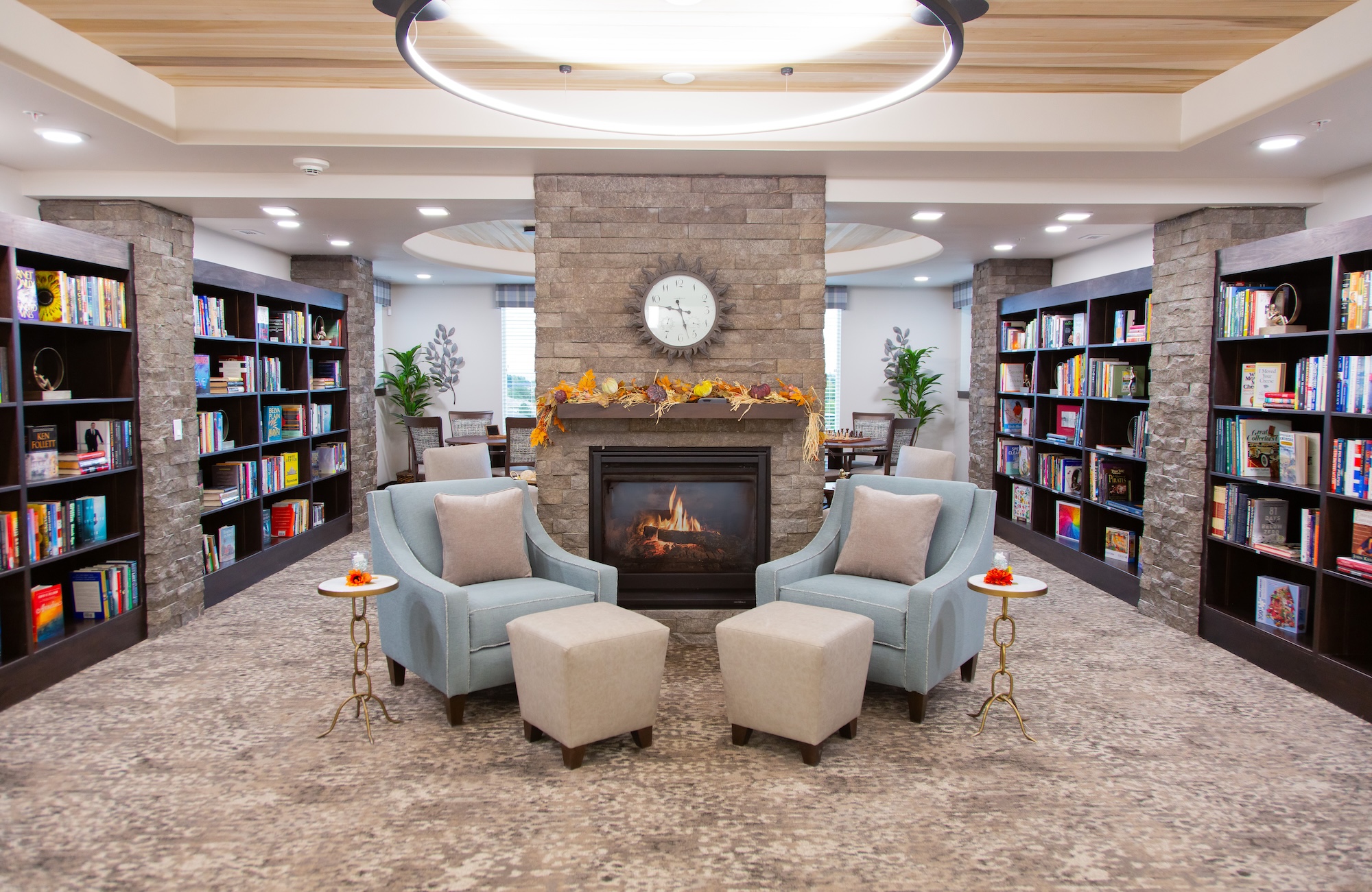 A cozy library with a stone fireplace, surrounded by bookshelves filled with books. Two blue chairs and two beige ottomans sit near the fireplace. A clock above the mantle and autumn decorations add warmth to the inviting space.