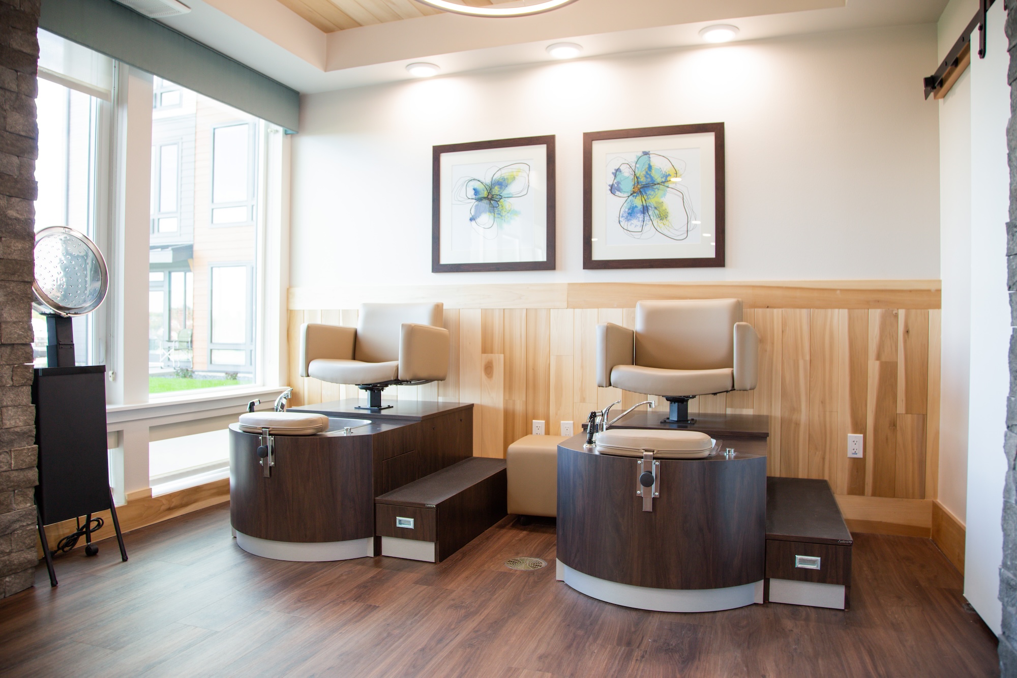 A modern nail salon interior with two pedicure stations featuring cushioned chairs and foot baths. The room has wooden wall panels, large windows, and two abstract floral paintings on the wall.