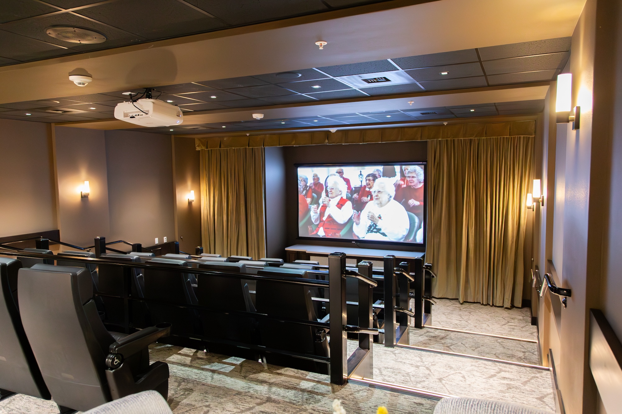 A home theater with multiple black leather recliners facing a large screen displaying a group of people. The room has dim lighting, beige walls, and a carpeted floor, with recessed lights and a ceiling-mounted projector.