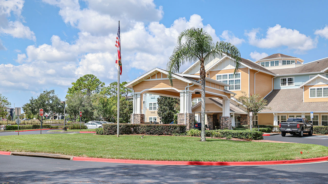 A two-story building with a covered entrance and stone pillars, surrounded by green lawns and palm trees. An American flag is on a flagpole near the entrance, and cars are parked nearby under a bright blue sky.