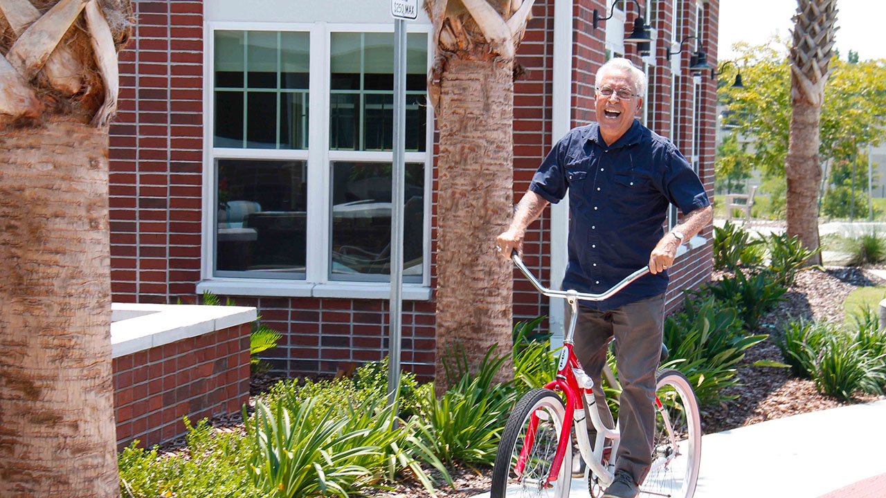 An older man with gray hair, wearing glasses and a navy shirt, smiles and rides a bicycle on a sidewalk next to a brick building and palm trees.