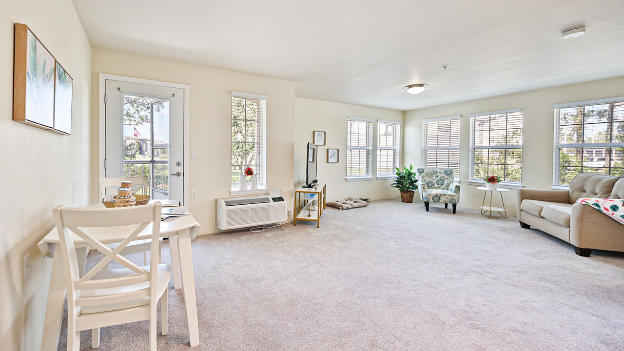 Bright, spacious living room with large windows, beige carpet, a desk with chairs on the left, a TV on a stand, a beige sofa, a patterned armchair, and a small dog bed by the wall.