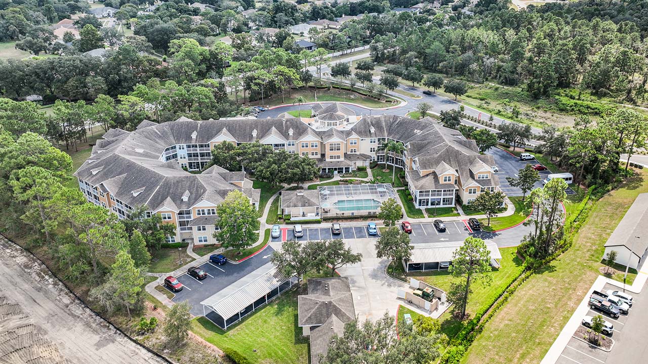 Aerial view of a large, multi-story senior living facility surrounded by trees and greenery, with a central courtyard, parking lot, covered carports, and nearby residential areas.