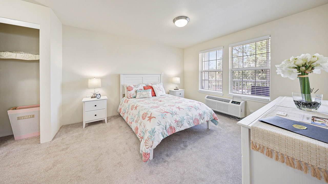 A bright bedroom with a white bed, floral bedding, two nightstands with lamps, a dresser with flowers, and large windows allowing natural light to fill the carpeted space. An open closet is visible on the left.