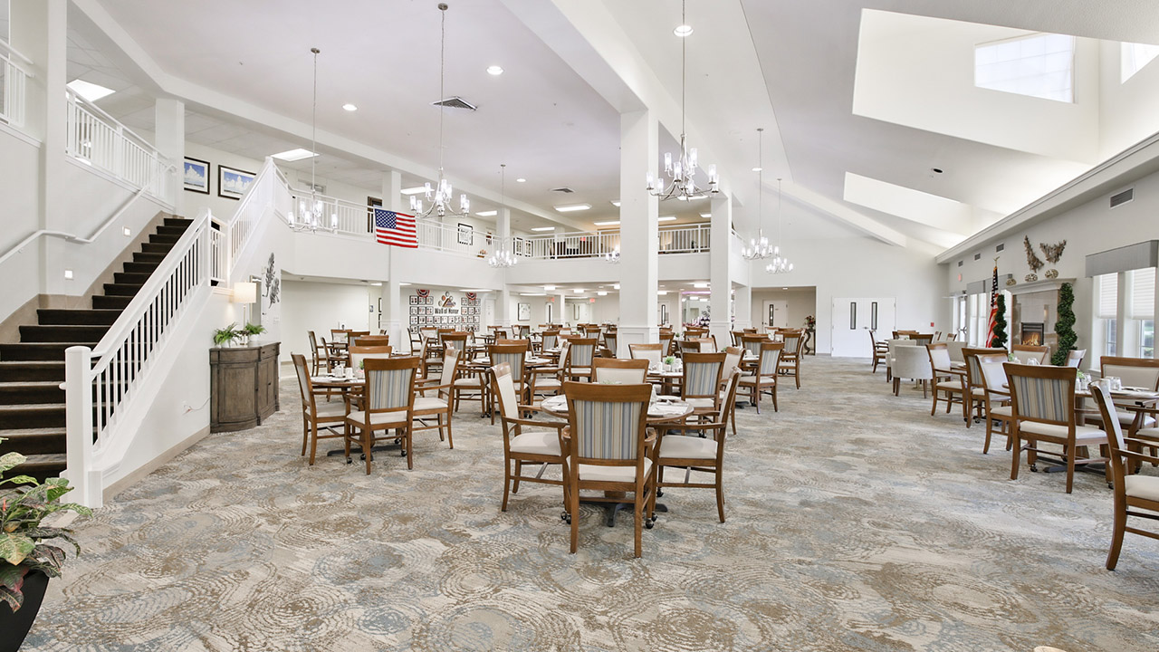 A spacious, well-lit dining area with neatly arranged wooden tables and chairs, chandeliers, an American flag, carpeted floors, and large windows letting in natural light. A staircase leads to an upper level.