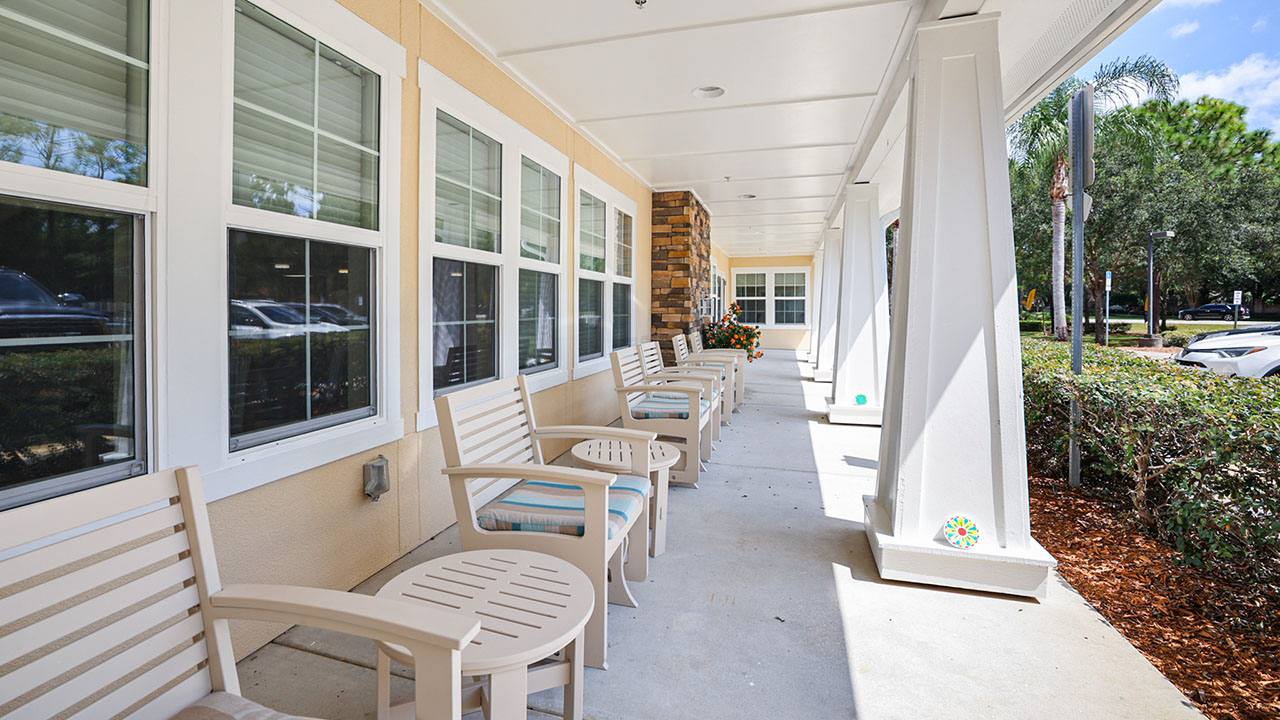A covered outdoor patio with several beige chairs and small round tables lined up along a row of windows, overlooking a landscaped parking area and greenery.