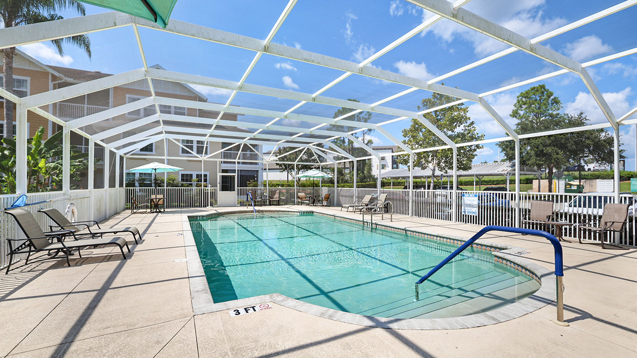 A covered outdoor swimming pool with clear blue water, surrounded by lounge chairs and tables with umbrellas, enclosed by a white frame and fence under a sunny sky.