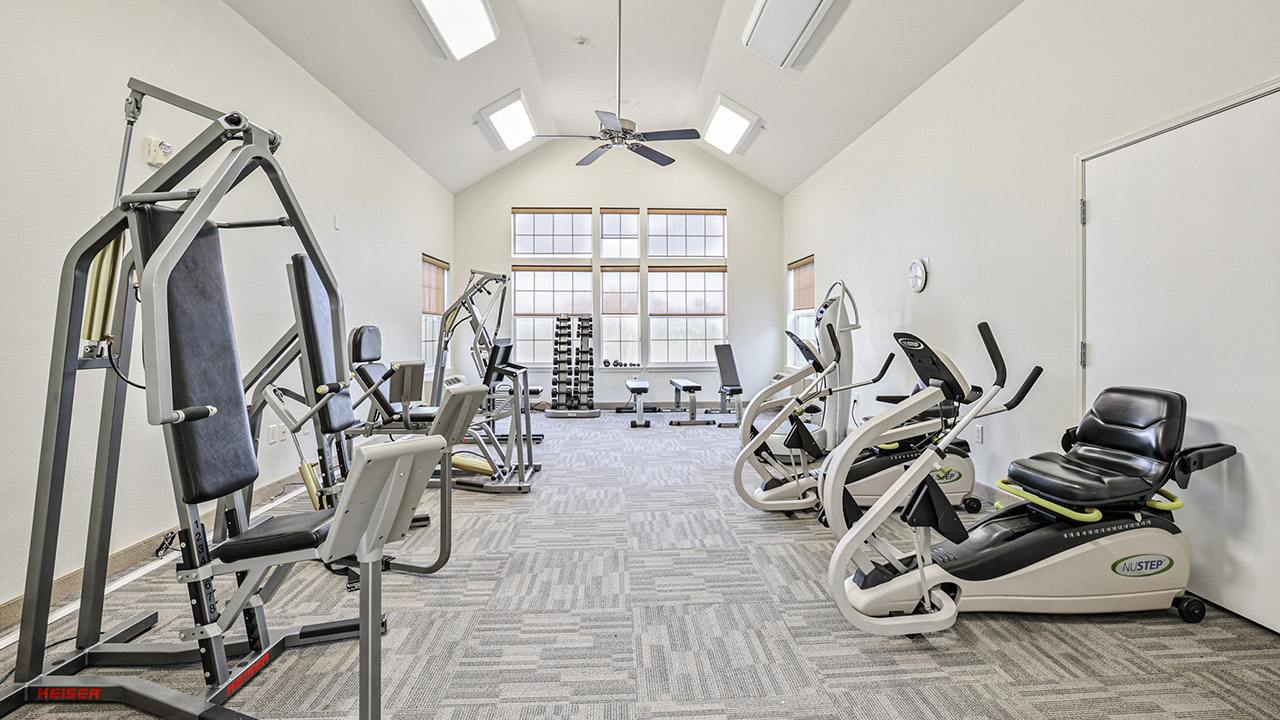 A bright, spacious gym room with various exercise machines including ellipticals, a multi-gym station, and free weights. Large windows let in natural light, and the room has light-colored walls and carpeted flooring.
