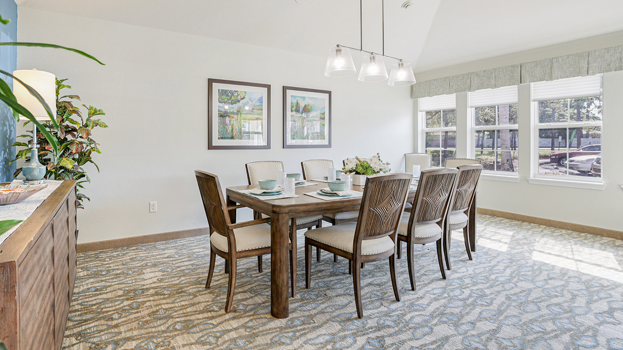 Bright dining room with a wooden table set for eight, cushioned chairs, large windows letting in natural light, two framed artworks on the wall, and a sideboard with plants and decor.