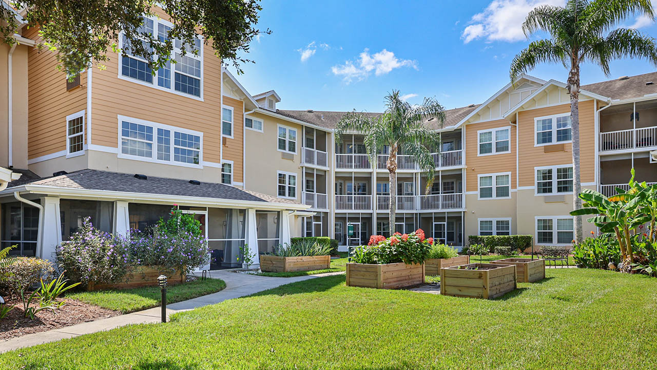 A well-maintained apartment complex with tan siding, white trim, and large windows. The courtyard features green lawns, raised garden beds, palm trees, and a clear blue sky overhead.