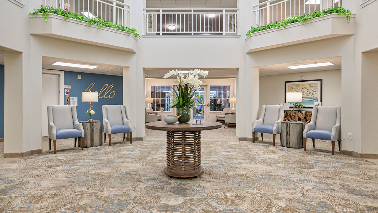 A bright, welcoming lobby with a round table and flower arrangement in the center, surrounded by blue cushioned chairs, side tables, and greenery on a balcony above. The walls are decorated with art and the word "hello".