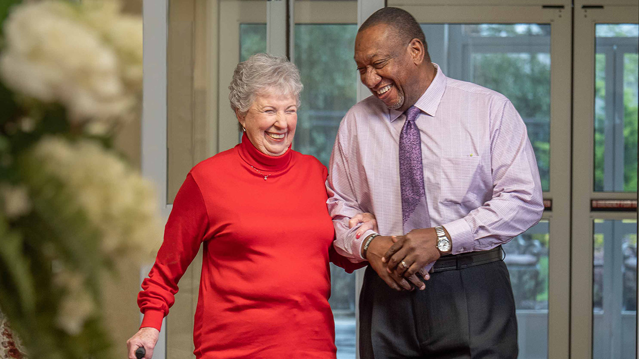 An elderly woman in a red turtleneck shirt smiles as she walks arm-in-arm with a man in a light purple dress shirt. They appear happy and are indoors near large windows.
