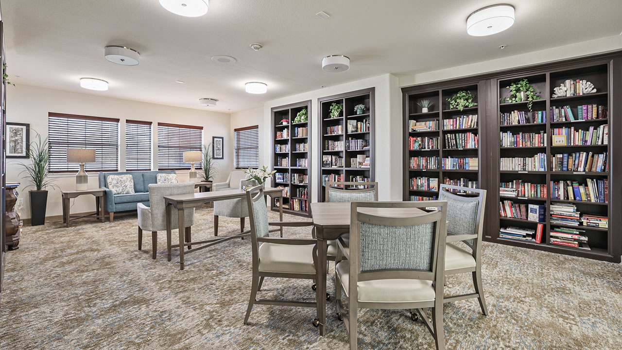 A spacious library room with bookshelves filled with books, several tables and chairs, armchairs near the windows, and decorative plants on the shelves and tables. Large windows let in natural light.