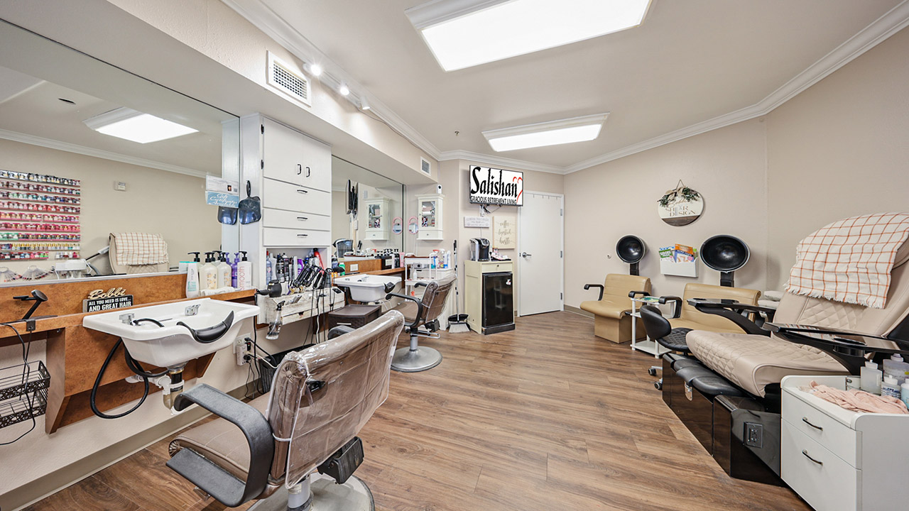 A bright, clean salon with barber chairs, hair washing stations, manicure tables, and two pedicure spa chairs, all on a wood floor. Shelves hold nail polish and supplies, and a mirror reflects the well-lit space.