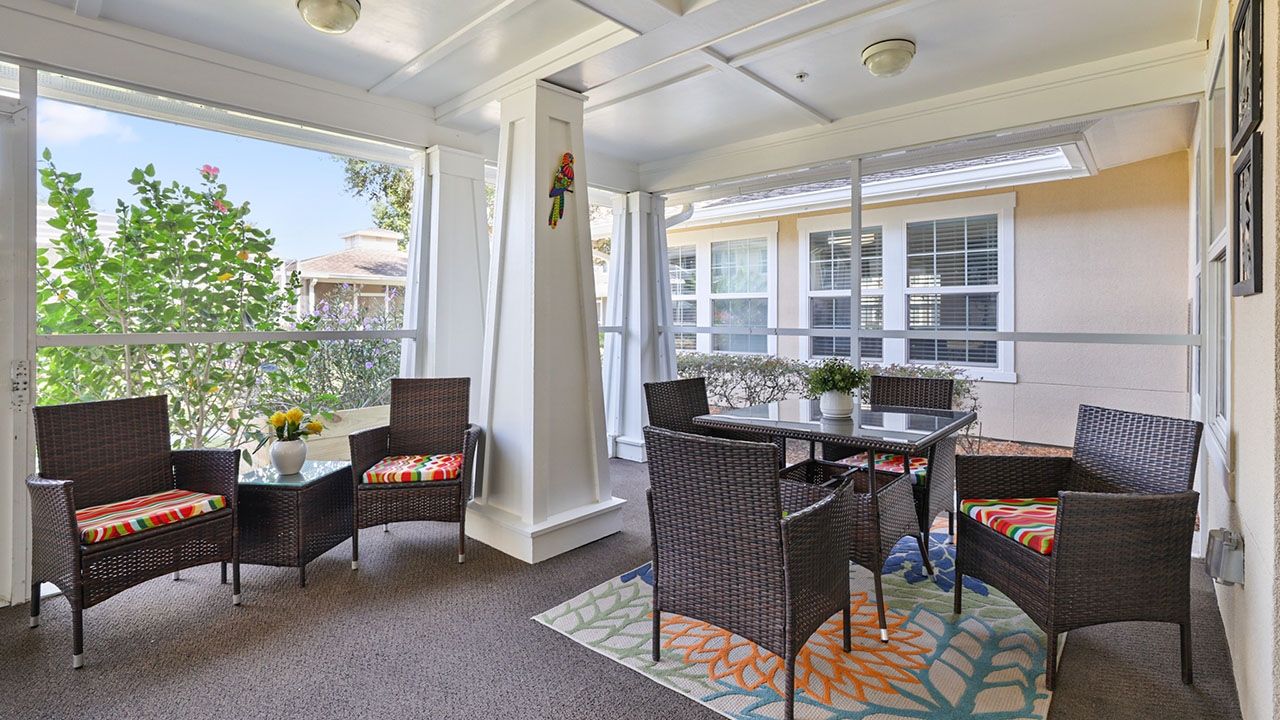 A bright sunroom with wicker furniture, colorful cushions, a glass-top table, potted plants, and large screened windows overlooking a garden and nearby houses.