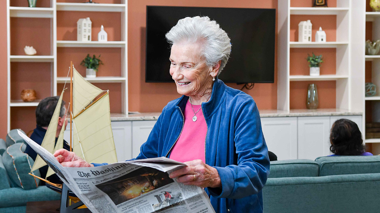 An elderly woman with short gray hair, wearing a blue jacket and pink top, smiles while reading a newspaper in a cozy living room with a large TV, shelves, and other people seated in the background.