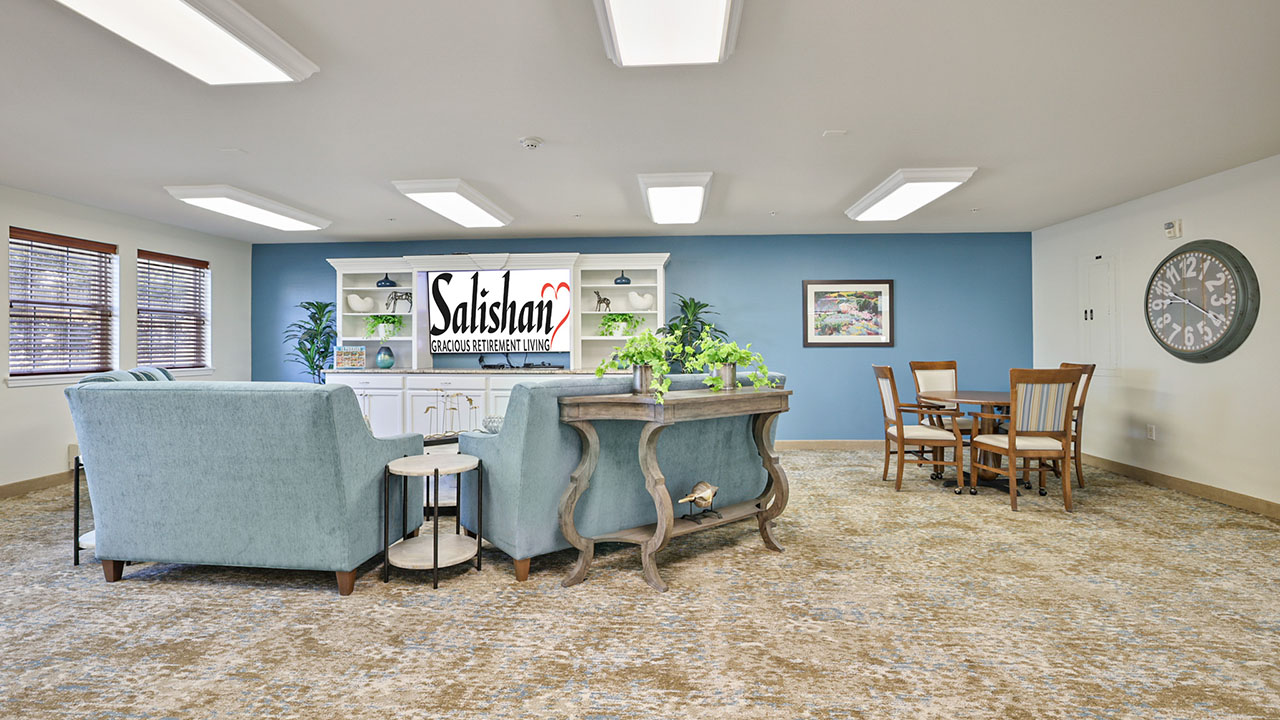 A bright, spacious living area with blue sofas, a wooden table with chairs, large wall clock, plants, and a blue accent wall featuring a sign that reads “Salisian SENIOR ASSISTED LIVING.”.