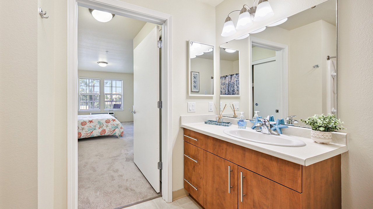 Modern bathroom with a wooden vanity, white countertop, and mirror, connected to a bedroom with beige carpet, a bed, and large windows letting in natural light.