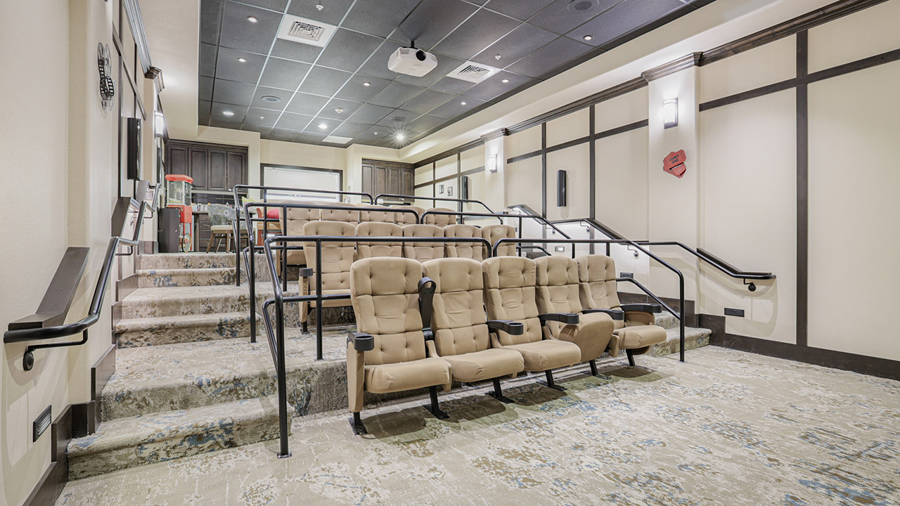 A small, modern theater room with three tiered rows of tan, cushioned seats, black railings, patterned carpeting, and wall-mounted lights, facing a projection screen at the front.