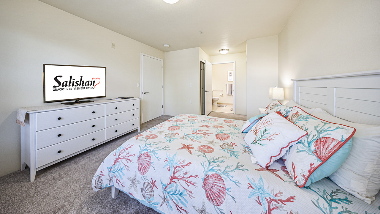 A bright bedroom with a coastal-themed bedspread, white furniture, a dresser with a TV displaying "Salishan," and an open door leading to a hallway and bathroom. The room has beige walls and carpet.