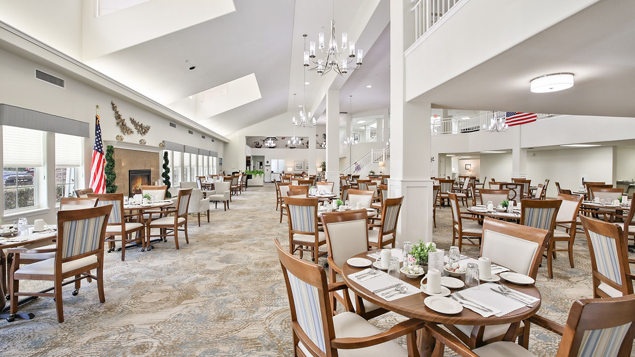 A spacious, well-lit dining room with neatly set round tables and wooden chairs, large windows, chandeliers, and American flags. The room has a modern, welcoming atmosphere with high ceilings and neutral decor.