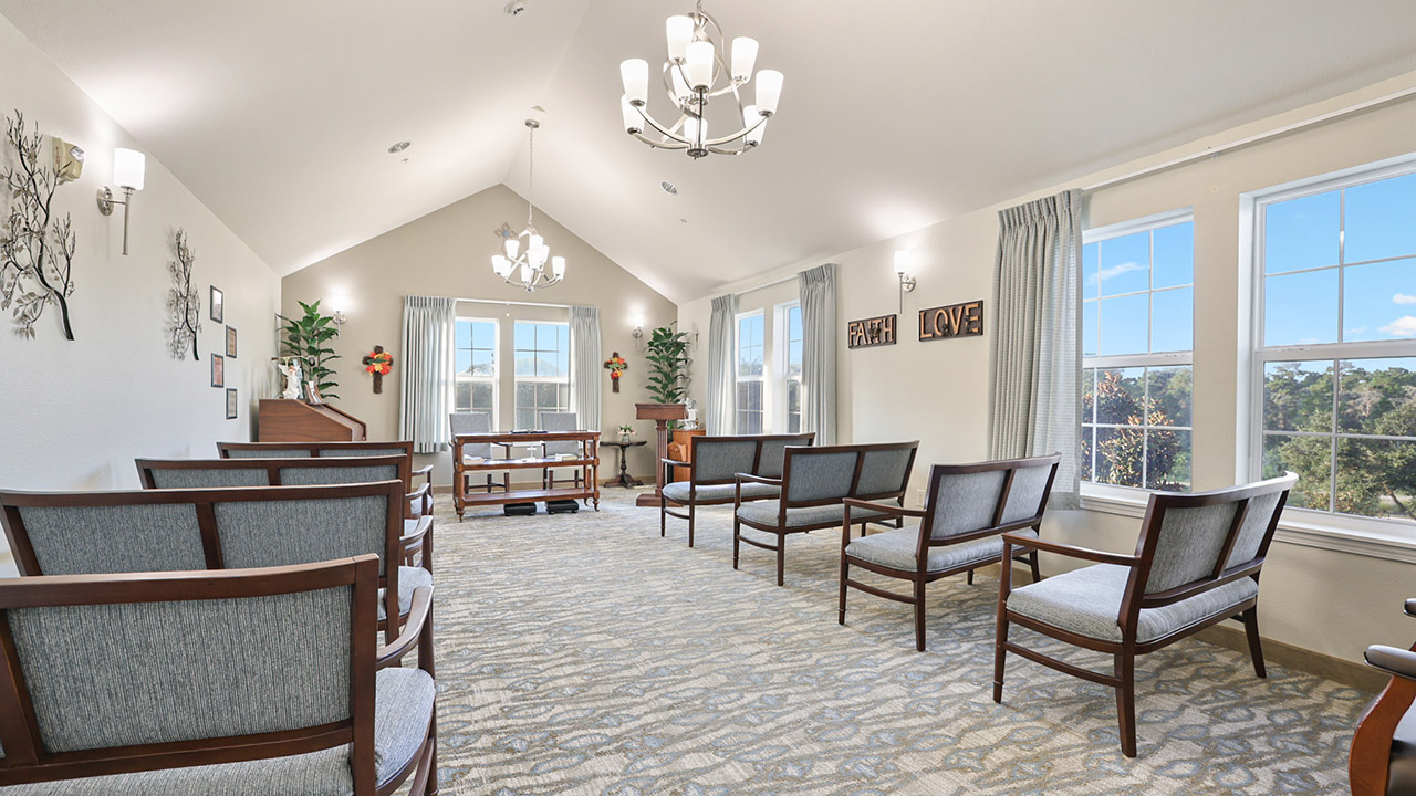 A bright, neatly arranged chapel with wooden chairs, large windows letting in daylight, chandeliers, and decor including crosses, plants, and wall art with the words “FAITH” and “LOVE.”.