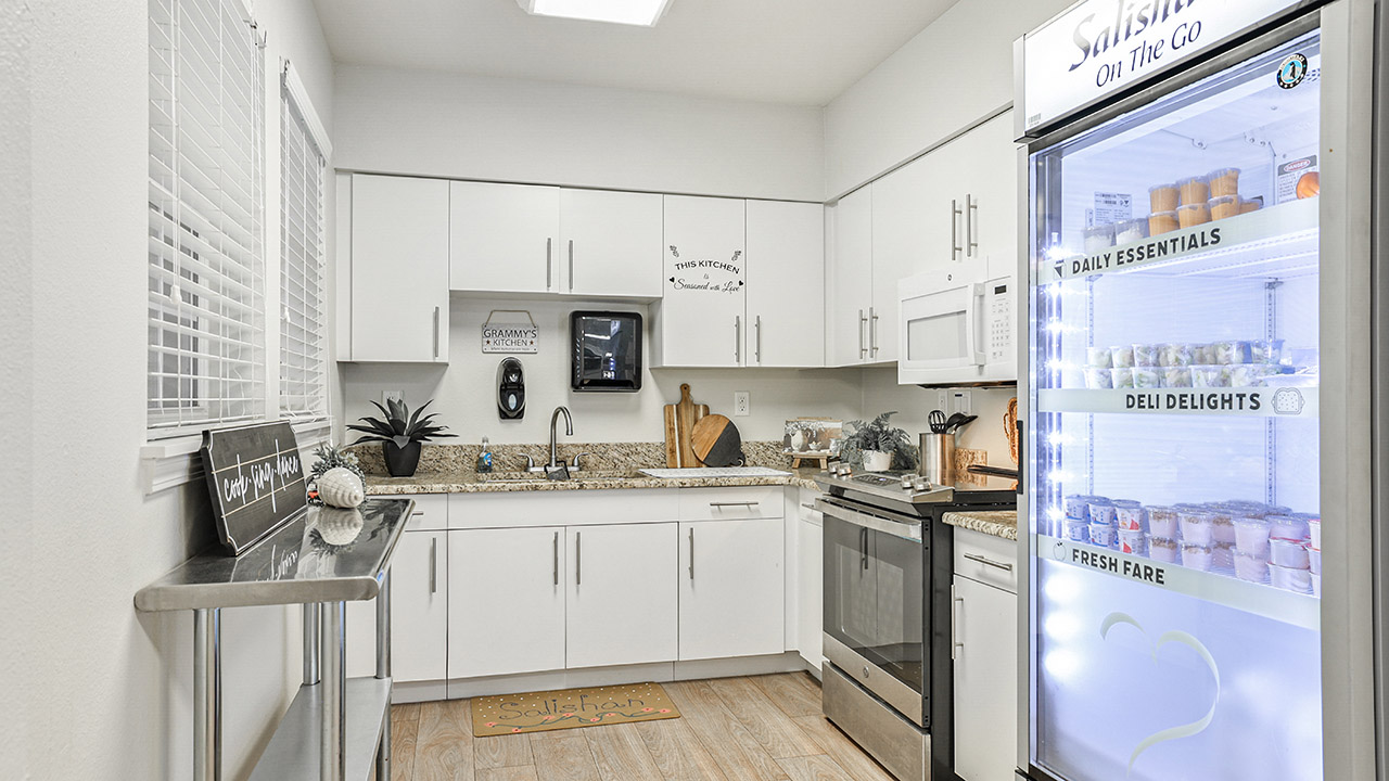 A modern, bright kitchenette with white cabinets, granite countertops, stainless steel appliances, and a glass-door refrigerator stocked with packaged food and drinks. A small plant and kitchen decor sit on the counter.