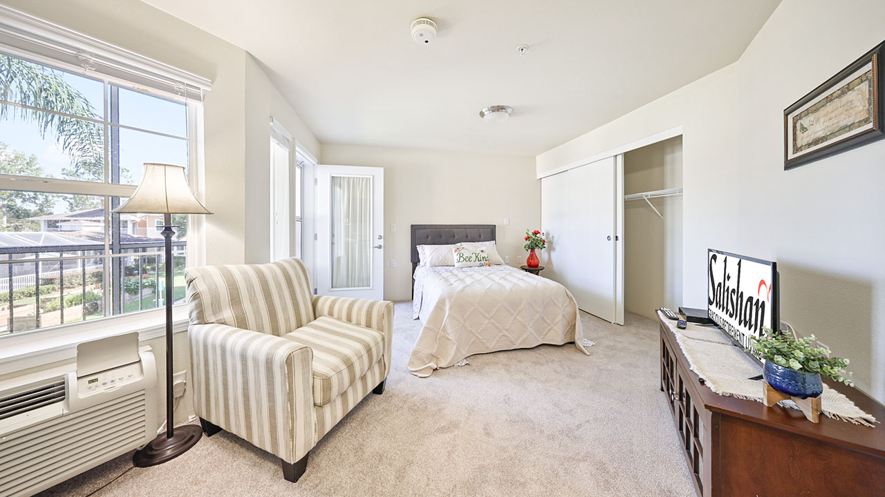 Bright bedroom with a bed, armchair, dresser with TV, and open closet. Large window and glass doors let in natural light. Room is decorated in neutral tones with a cozy, inviting atmosphere.