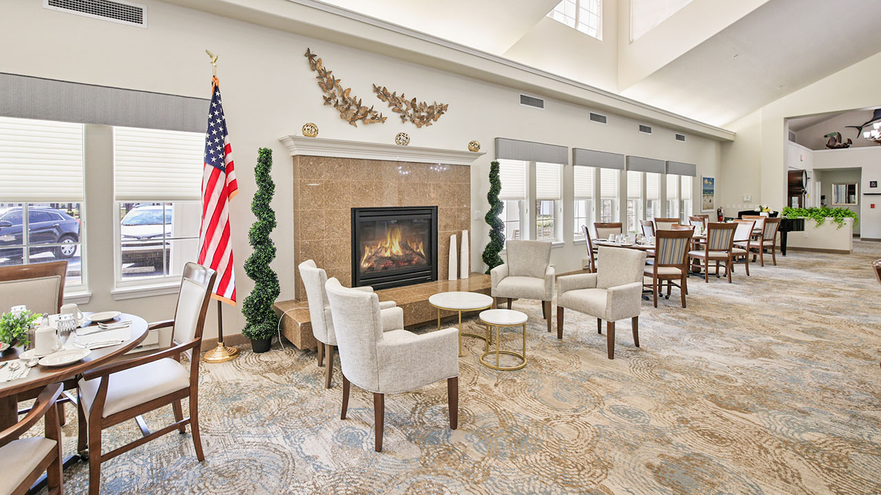 A bright, spacious dining area with beige armchairs and tables arranged near a fireplace. An American flag stands by the hearth, and large windows let in natural light. Decorative branches hang above the mantel.