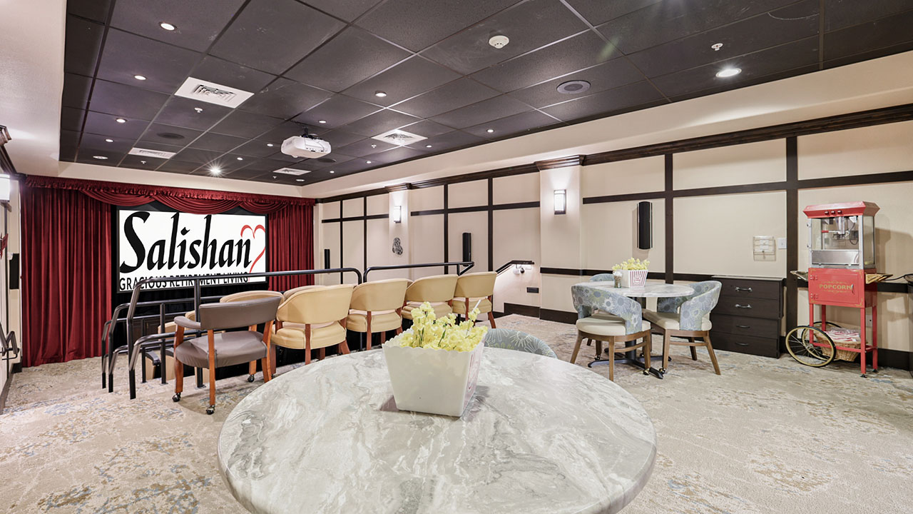 A small, modern home theater room with a movie screen labeled "Salishan," rows of chairs, round tables with flowers, soft lighting, and a popcorn machine in the corner.