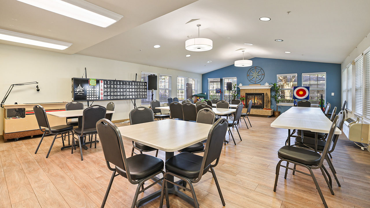 A bright, spacious community room with tables and chairs arranged for group activities. There is a fireplace, bingo board, air hockey table, and large windows letting in natural light.
