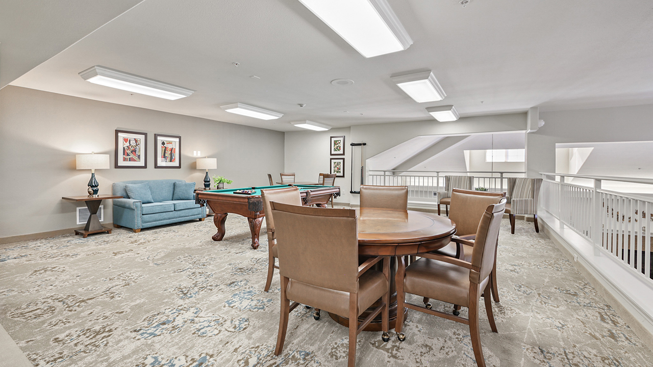 A spacious, well-lit game room with a pool table, a blue sofa, framed art on beige walls, a round table with four brown chairs, and large windows with white railings. The floor is covered with a patterned rug.