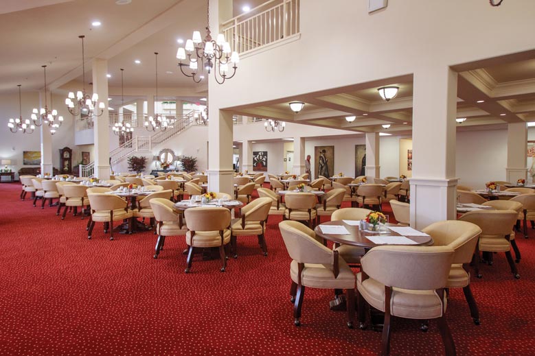 A spacious dining area with beige chairs and set tables, featuring a red carpet and elegant chandeliers. The room has high ceilings, large windows, and a staircase leading to an upper level. Decorative plants and artwork enhance the warm, inviting atmosphere.