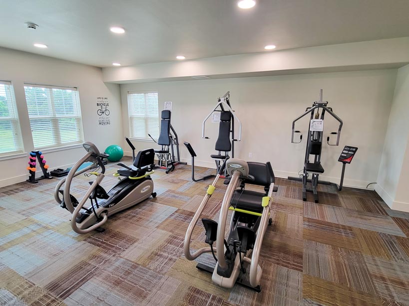 A small gym room with exercise machines, including an elliptical trainer, a stationary bike, a cable machine, and a seated leg press. Free weights and a green exercise ball are near the window in the corner. The room is well-lit by several windows.