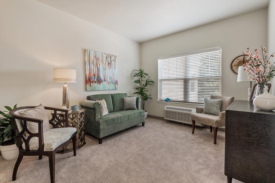 A cozy living room featuring a green two-seater sofa with two light-colored cushions, flanked by two wooden chairs with cushion seating. A coffee table sits adjacent to the sofa. Decor includes plants, a tall lamp, artwork, a window with blinds, and a sideboard.