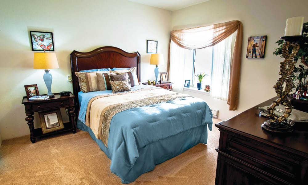 A cozy bedroom with a large bed featuring a blue comforter and multiple decorative pillows. The room includes wooden furniture, such as two side tables with lamps, a dresser with a mirror, and a window adorned with sheer brown curtains. Framed pictures decorate the walls.