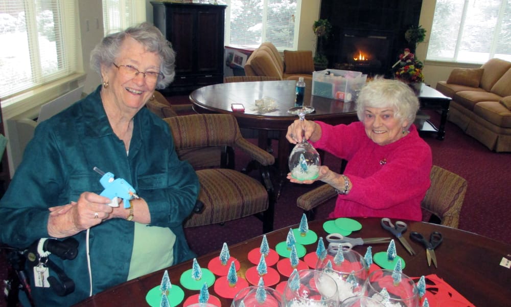 Two elderly women, seated at a table, are crafting holiday decorations using wine glasses. The woman on the left is smiling and holding a hot glue gun, while the woman on the right displays a decorated glass. The table is covered with materials and finished pieces.