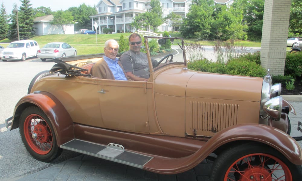 Two men sitting in a vintage tan and brown convertible car with red spoked wheels. The older man on the left wears a brown jacket, and the younger man on the right wears a gray t-shirt and sunglasses. Residential buildings and cars are visible in the background.