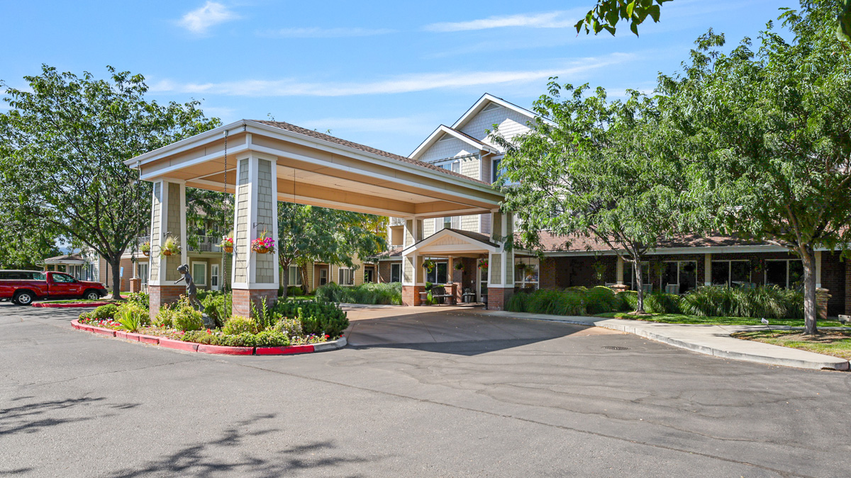 A large, covered entrance with columns leads to a modern, multi-story building surrounded by trees, neatly trimmed bushes, and a paved driveway on a sunny day.