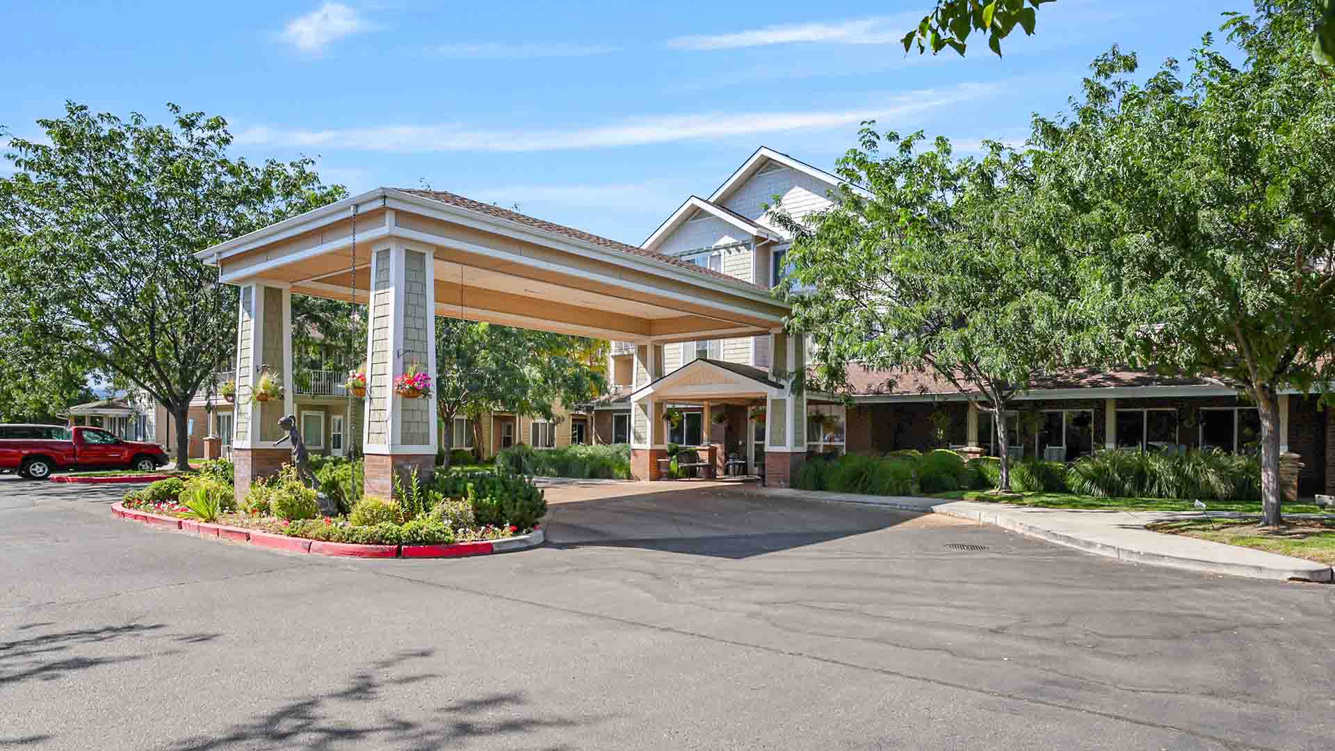 A large building with a covered driveway entrance, surrounded by trees, greenery, and a parked red vehicle, under a blue sky with light clouds.
