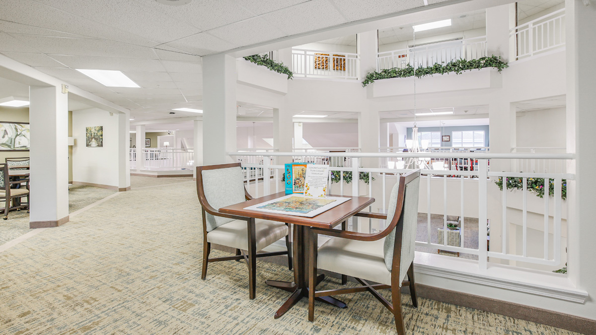A small table with two chairs is set up near a white railing on an upper floor of a bright, open indoor atrium. The table holds puzzle pieces and board games. The area has neutral colors and green plants along the railing.