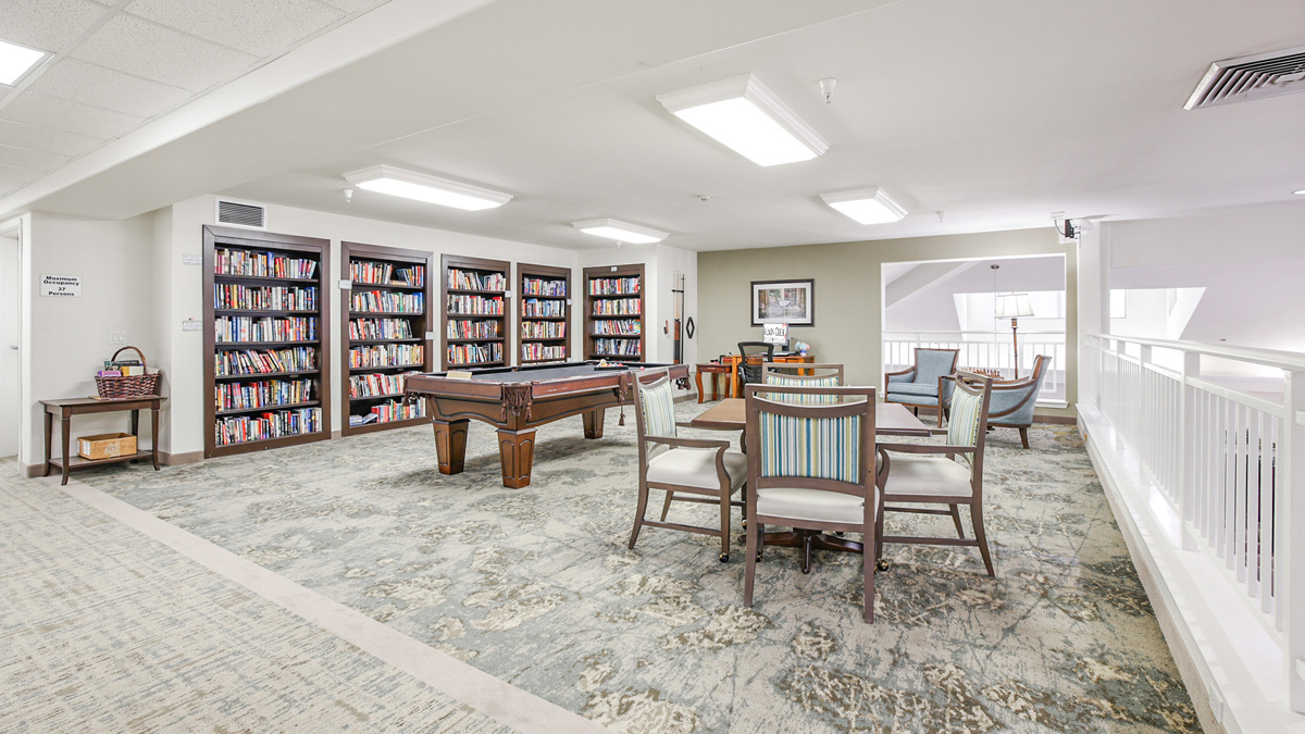 A bright, open library room with bookshelves along one wall, a pool table in the center, several tables and chairs, and carpeted floors, featuring modern lighting and a railing overlooking another level.