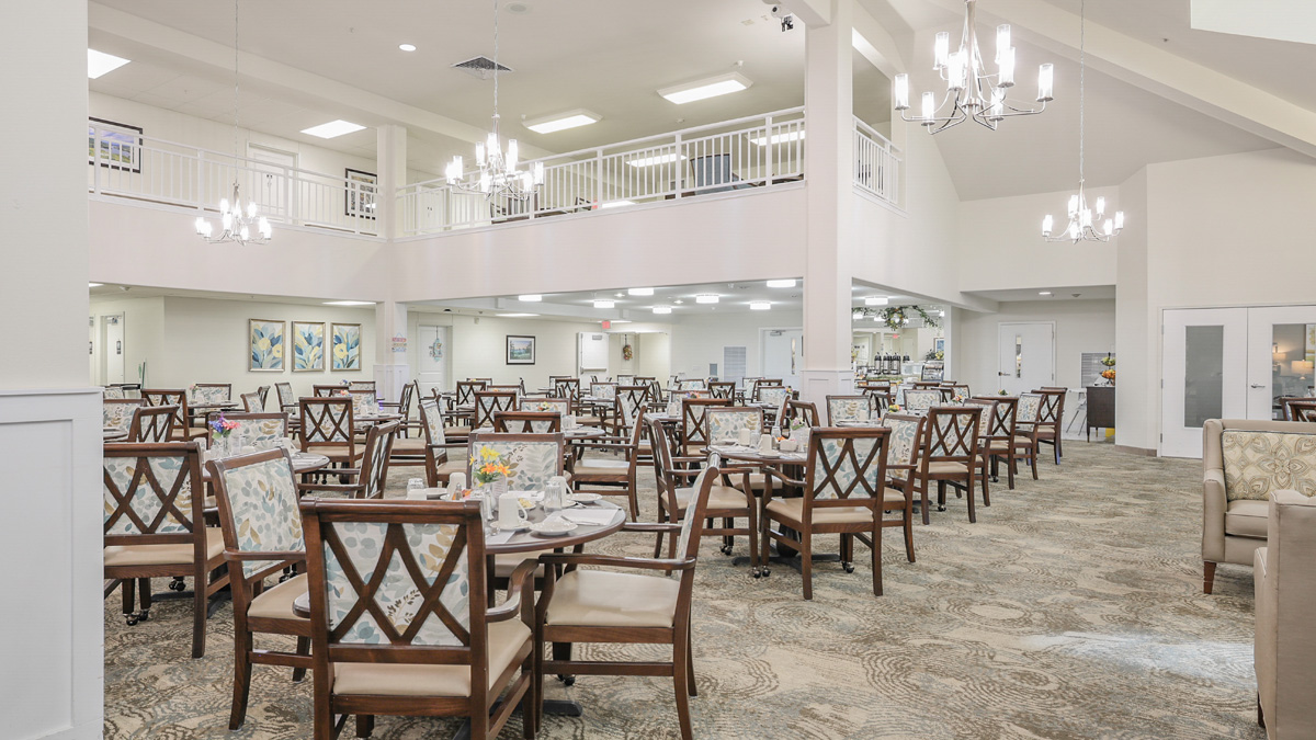 A spacious, well-lit dining room with many round tables and wooden chairs, set for meals. Chandeliers hang from the ceiling, and there is a balcony above. The decor is light and elegant, with paintings on the walls.
