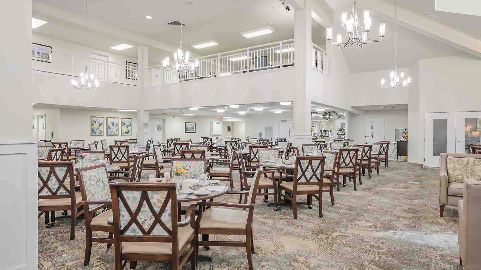 Bright, spacious dining area with multiple round tables and wooden chairs, elegant chandeliers, patterned carpet, and artwork on the walls; room has high ceilings and an upper balcony.