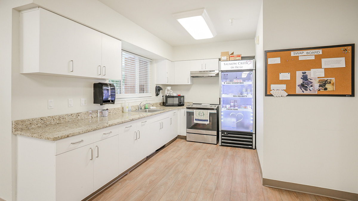 A clean, modern kitchen with white cabinets, a granite countertop, and wooden floor. There is a refrigerator, microwave, stove, and a corkboard labeled “SWAP BOARD” on the wall. A window lets in natural light.