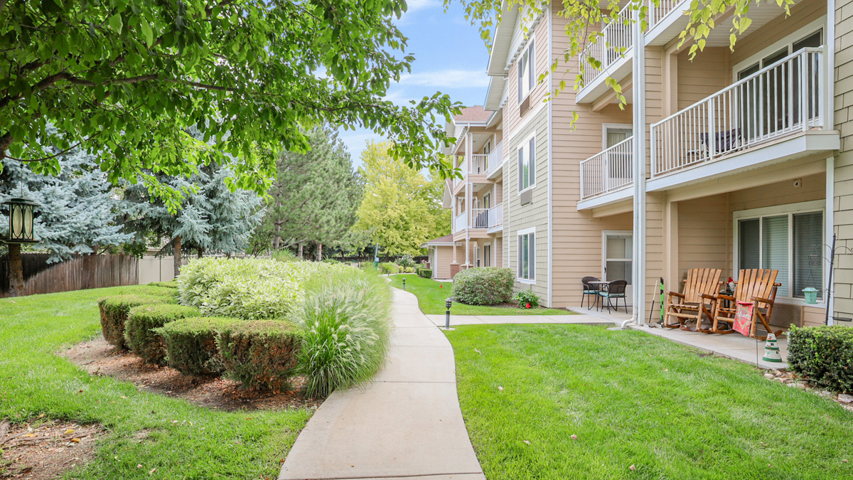 A paved walkway winds through a well-kept lawn with shrubs and trees, alongside a beige three-story apartment building with balconies and patio furniture.