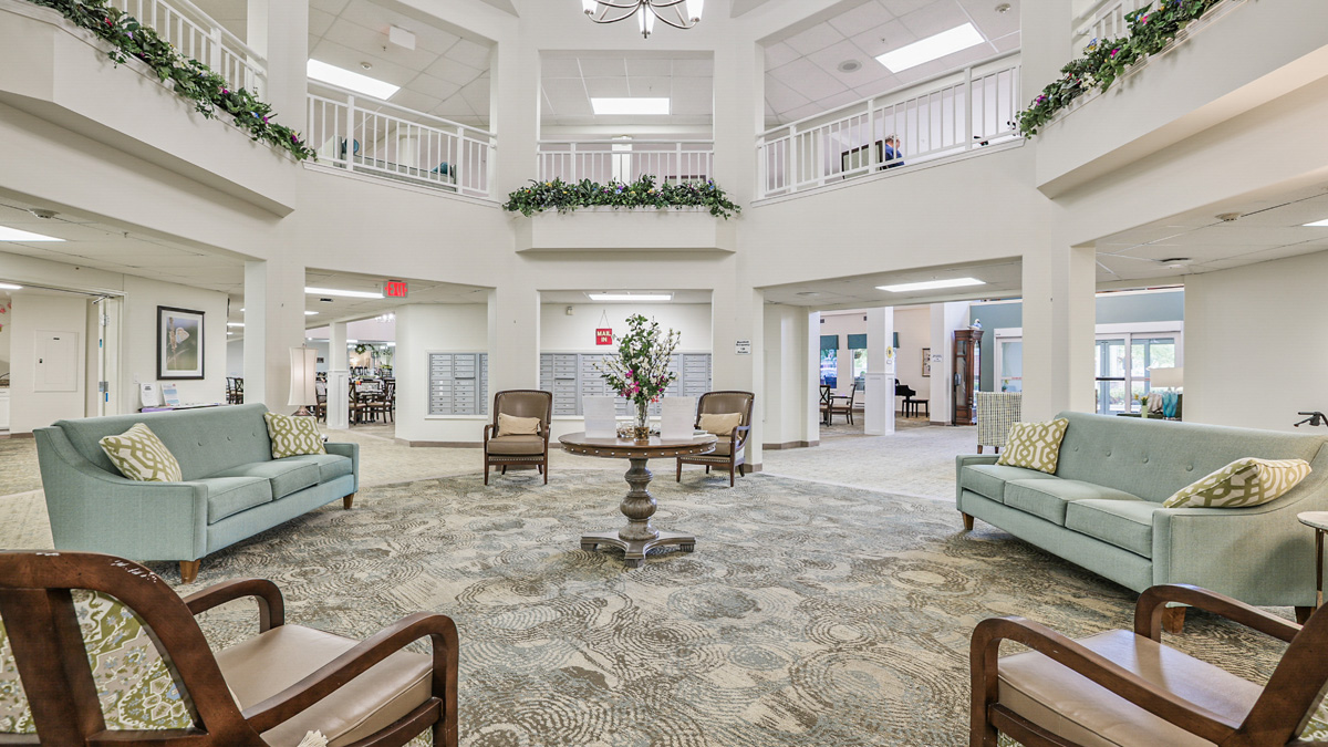 A spacious, well-lit lobby with two light blue sofas, four wooden chairs, a round table with a flower arrangement, and greenery on the upper railings under a high ceiling.