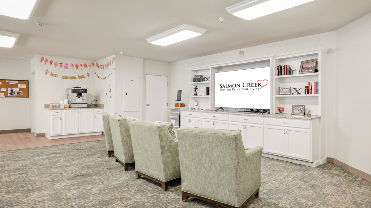 A cozy room with four green armchairs facing a TV displaying "Salmon Creek Gracious Retirement Living," white cabinets, bookshelves, a coffee station, and cheerful decorations hanging from the ceiling.